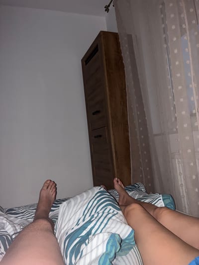 The image shows a person's legs and feet in a bed. The person is lying on their side, with their legs bent. The bed has striped bedding in blue, white, and green. There is a wooden shelf mounted on the wall above the bed, with two dark drawer pulls. The scene appears to be indoors, possibly in a bedroom. The lighting is dim, suggesting it may be nighttime or early morning. There is no visible text in the image. The overall mood is relaxed and intimate. The location context provided is Toruń, Poland, but there are no specific visual cues to confirm this.