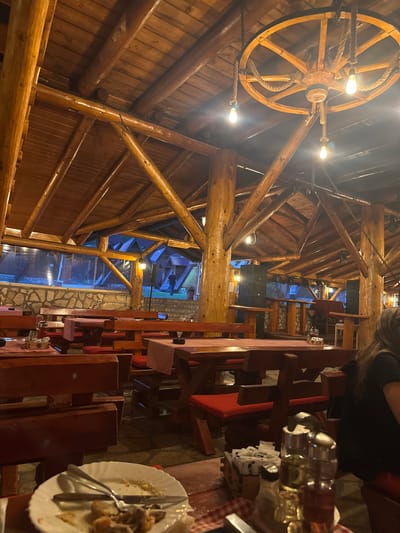 The image shows an outdoor dining area of a restaurant, likely in Bastasi, Bosnia and Herzegovina, characterized by its rustic, wooden architecture. The ceiling is constructed from thick, natural logs and wooden planks, supporting a sloped roof. A large, decorative wooden wagon wheel hangs from the ceiling, serving as a chandelier with exposed light bulbs.

The seating consists of sturdy, wooden picnic tables with benches, many of which have red cushions. Red and white checkered tablecloths cover some of the tables, and condiments are visible, including salt and pepper shakers and what appear to be oil and vinegar bottles. In the foreground, a plate with leftover food and cutlery is on a table.

In the background, several small, triangular-roofed structures, resembling cabins or chalets, are visible, illuminated by warm lights. A person is seen walking in the distance between these structures. The overall ambiance suggests a casual, outdoor dining setting, possibly a lodge or a traditional Bosnian restaurant. The lighting is warm and ambient, indicative of evening or night.