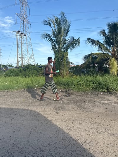 A young Black male, walking left-to-right across an unpaved, dusty path in Ojomu, Nigeria, is the central figure. He wears a dark sleeveless shirt with a faded graphic, vertically striped green and white trousers, and dark flip-flops. A dark backpack is visible on his back. He holds a light green container in his left hand, while his right hand is raised to his mouth. Overgrown green grass and bushes line the path behind him, with additional vegetation and indistinct structures in the mid-background. To the left, a tall metal lattice power transmission tower stands against a clear blue sky with sparse white clouds. Multiple power lines traverse the sky. Several palm trees are visible on both the left and right sides of the frame, some partially obscured by power lines. The ground is a mix of dirt and gravel, with a shadow cast across the foreground from an unseen source to the right.