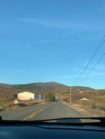 The image, taken from within a vehicle, captures a mountainous landscape under a bright, clear blue sky. A paved road with double yellow lines extends into the distance, appearing to ascend gently through the terrain. The right side of the frame is obscured by a dark shape, likely the interior of a car's window frame or pillar.

To the left of the road, the ground is covered in dry, sparse, brownish vegetation and scattered trees, suggesting an arid environment or the autumn/winter season. A prominent green coniferous tree stands out near a cluster of modern buildings. One building features white walls and a light peach or pink-colored roof, while an adjacent structure has a roof covered in dark grey or black panels, possibly indicating solar installations. A small, triangular road sign with a red border is visible near these buildings, though its details are indistinct.

Further into the background, a large, dark mountain or hill range dominates the horizon beneath the expansive blue sky. Overhead, several power lines stretch across the sky, supported by wooden utility poles lining the road. The sky is largely clear, with only a few faint, wispy clouds.

The scene depicts a journey through a rural or semi-rural mountainous region, consistent with the provided context of Grevena, Greece. The bright, direct sunlight suggests it is midday or early afternoon on a clear, sunny day. No people or specific activities are visible, and no legible text appears in the image. The overall impression is one of natural beauty and serene travel.