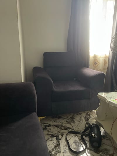 The image depicts an indoor scene, likely a living room, with two dark-colored sofas. One sofa is partially visible in the foreground, and a larger, single-seater armchair is prominent in the middle ground. The armchair is upholstered in a dark fabric and has a plush appearance. Behind the armchair, a window is covered by sheer curtains, allowing bright sunlight to filter through, creating a luminous effect. The window has decorative elements at the bottom. To the right of the armchair, the edge of a fan is visible. The floor is covered in marble-patterned tiles. On the floor near the armchair, a black dog leash with a metal clip is lying on the tiles. A portion of a white piece of furniture, possibly a side table, is visible on the right edge of the image. The overall lighting suggests it is daytime. The location is identified as Naze, Nigeria.