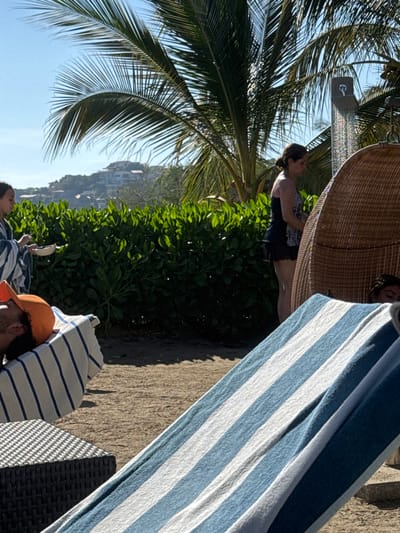 A beach scene shows a woman showering under a palm tree. Next to her is a wicker chair with someone inside. In the foreground, a blue and white striped towel is draped over a chair, with a man in an orange hat resting beneath it. Lush greenery and buildings on a hill fill the middle ground. The focus is sharpest on the figures and foreground elements, with the background slightly blurred.