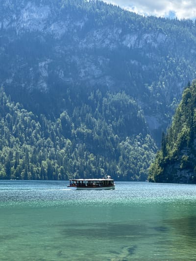 This image captures a serene view of Königssee lake in Schönau am Königssee, Germany. A white and brown passenger boat with several people visible inside is navigating the crystal-clear, turquoise waters. The boat is moving from left to right, leaving a gentle wake. The lake is surrounded by steep, densely forested mountains that rise dramatically in the background. The trees cover the mountain slopes from the water's edge to the rocky peaks. The sky above is partly cloudy. The overall scene conveys a sense of tranquility and natural beauty.