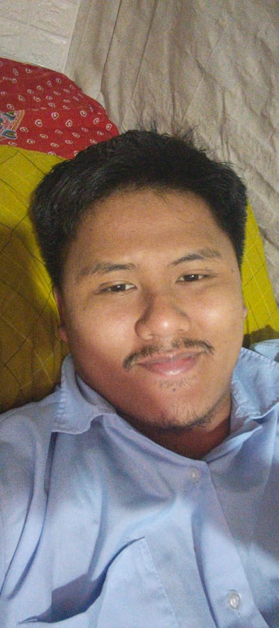 A close-up selfie taken from a low angle shows a person's face and upper chest, positioned in the foreground. The subject, appearing to be male, has dark hair, a mustache, and a small amount of stubble on his chin. He is wearing a light blue collared shirt. The background consists of a wrinkled, light-colored fabric, a section of a red fabric with a white circular pattern, and a yellow surface with a grid-like texture. There are no discernible city landmarks or specific environmental cues to indicate a location in Kemuning, Indonesia. The lighting is dim, and the image has a grainy quality.