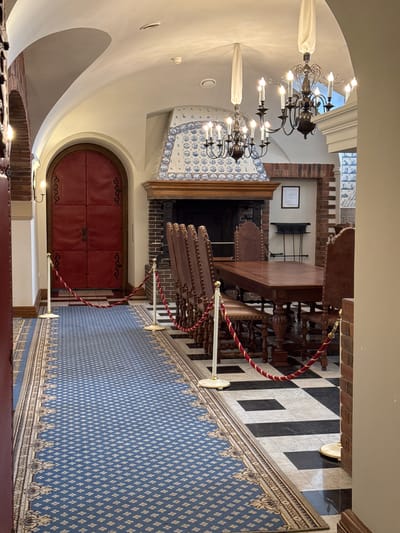 The image shows an interior view of a hallway and dining area, likely within a historical building. A large, arched red door with decorative metalwork is visible on the left. A blue patterned carpet with an ornate border runs down the hallway, bordered by stanchions with red and gold ropes. To the right, a long wooden table is set with multiple chairs, suggesting a dining room. A prominent fireplace with blue and white patterned tiles sits behind the table. Two ornate chandeliers hang from the arched ceiling. The floor in front of the fireplace and dining area is tiled in a black and white checkered pattern. The overall aesthetic suggests a formal and possibly historical setting. Based on the architectural style and the hint that the location is in Strelna, Russia, this is likely part of the Constantine Palace or a related building within the palace complex. No people are present in the image.