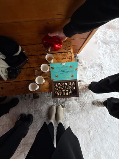 The image captures an outdoor winter scene in Yeghipatrush, Armenia. A dark-paneled wooden surface, possibly a bench or table, occupies the upper portion of the frame. On this surface, a hand clad in a dark sleeve is pouring a light-colored liquid from a red, irregularly-shaped bottle into one of four white paper cups. Three additional empty white paper cups and a small, clear, textured glass bottle are also present. An open box of "MILLENNIUM 16 SWEETS MILK CHOCOLATE COLLECTION OCEAN STORY" is displayed, revealing a selection of marine-themed chocolates arranged within a dark tray. To the left of the wooden surface, several dark and light articles of clothing are stacked. The ground below and extending to the right is covered in an uneven layer of snow and ice. The lower extremities of three individuals are visible: a pair of feet wearing black pants and light-colored sneakers (right), a pair in dark striped pants and black chunky boots (left), and a pair wearing a long black coat and light-colored fur-lined boots (foreground).