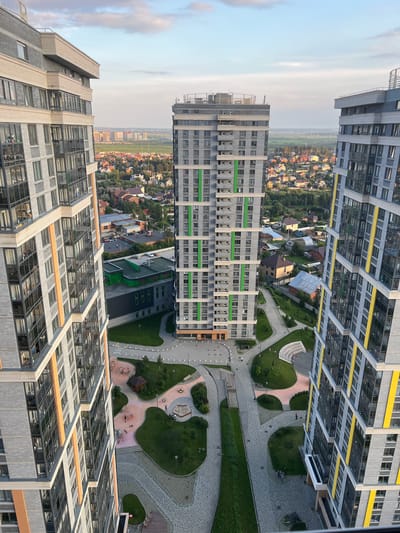 This is an aerial view of a residential complex in Sovkhozny, Russia, likely taken from a high floor of one of the apartment buildings. The scene shows multiple modern high-rise apartment buildings with gray brick exteriors and large windows. Some buildings feature accent colors like green and yellow stripes. The buildings are arranged around a landscaped courtyard with pathways, green spaces, children's play areas, and seating benches. In the background, a suburban landscape with scattered houses and trees is visible, extending towards a distant city skyline. The sky is partly cloudy, with the soft light suggesting it is either early morning or late afternoon. Several individuals are visible on the ground, walking along pathways or in the courtyard, indicating active use of the communal space.