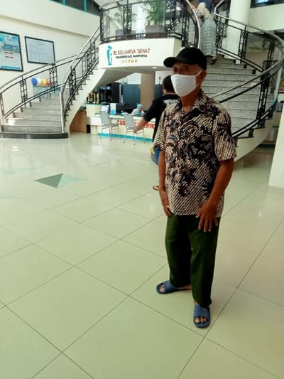The image shows a man standing in the lobby of a building located at coordinates -6.76 latitude and 111.015 longitude in Kecamatan Margorejo, Indonesia. The man is wearing a black cap, a patterned shirt, and green pants. He is wearing a white face mask. He is looking at the camera. The building has a tiled floor and a staircase with a railing. The background shows a sign reading "RS KELUARGA SEHAT TERAKREDITASI PARIPURNA," an information desk, and a person in a blue headscarf seated behind the desk. There is a man wearing a black shirt behind the first man. A woman in a long white dress is walking up the stairs. The image was taken inside the building.