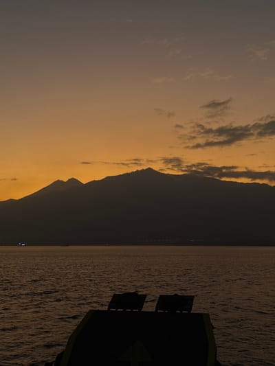 The image depicts a tranquil scene at dusk, likely near Gunung Malang, Indonesia. In the foreground, the dark silhouette of a ferry deck is visible, with two dark seating structures positioned at the rear. The sea stretches out before it, its surface rippling with a gentle, dark sheen. In the distance, a majestic mountain range forms a prominent, dark silhouette against the warm, gradient sky, transitioning from a deep orange near the horizon to a dusky grey higher up. Scattered clouds are visible, tinged with the fading light. On the horizon, distant lights suggest a coastal settlement or other vessels. The overall atmosphere is one of calm and serenity as day transitions into night.