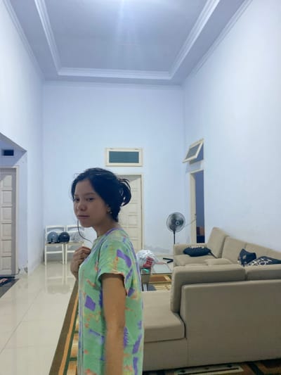 A young woman with dark hair pulled into a bun stands in what appears to be a living room. She is wearing a light green t-shirt with abstract purple and yellow patterns. She is looking off to her left, with her left hand near her neck and her right arm at her side. The room is brightly lit, with white walls and a high, ornate ceiling. In the background, there is a beige sectional sofa with several pillows, a standing fan, and two doors. A hallway leads off to the left, with another door visible. The floor is tiled with a patterned rug in the foreground. The overall impression is of a domestic indoor space. Without additional context, it is not possible to determine the specific location within Walennae, Indonesia.