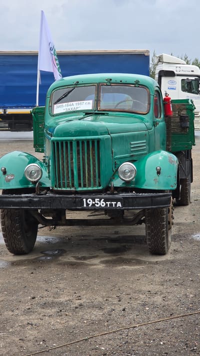 This media file depicts a vintage, bright green ZIL-164A truck from 1957, identified by text on the windshield and a license plate reading "19-56TTA". The truck is parked outdoors on a gravelly, wet surface, suggesting recent rain or a damp environment. In the background, a white KAMAZ truck with "Poctex" branding and a blue container are visible, hinting at a transportation or industrial setting, possibly a lot or depot. A white flag with text, partially obscured, is attached to the ZIL truck. The overall condition of the ZIL truck indicates it's well-maintained, possibly as a historical vehicle or for display, with some wear and tear evident on its paint. The lighting suggests it's daytime. The image is oriented vertically, with the truck appearing to be on its side.