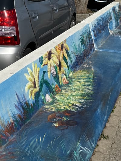 This media file captures an outdoor daytime scene, likely in Santo Stefano al Mare, Italy, featuring a long, low concrete structure that is adorned with a vibrant mural.

The mural depicts a lively natural landscape. Prominent yellow flowers, resembling sunflowers or large daisies, are painted across the surface, accompanied by smaller pink, orange, and white blossoms. Areas of lush green and yellow foliage, suggestive of tall grasses, reeds, and palm-like fronds, fill the background and foreground of the painted scene, all set against a vibrant blue sky or water. Two human figures, visible from behind, are integrated into the artwork. They appear to have dark skin, wear hats, and carry what look like baskets or bundles, seemingly moving through the painted fields or landscape. The painting exhibits visible brushstrokes, giving it an artistic style that combines elements of impressionism and folk art.

In the physical foreground and to the left, a dark grey or silver car is partially visible, showing its rear taillight and side body panels. Another darker vehicle is discernible further back. The ground surface around the painted structure is paved with grey interlocking concrete blocks, with small patches of weeds growing in the crevices. The bright illumination suggests it is mid-day with clear weather. No discernable text is visible within the image. The scene portrays a public space enhanced by the decorative, large-scale painting.