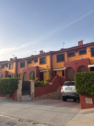 The image captures a row of attached houses in Urbanización Cabo Roig, Spain. The houses are painted in bright orange and deep red colors, with tiled roofs. Each house has dark-shuttered windows. In front of the houses, there are well-maintained green hedges and stone walls. A black metal gate is visible on the left side of the image. A silver Chevrolet Captiva SUV is parked in the driveway of one of the houses, facing away from the camera. The license plate reads "7470 LXK". The ground in front of the houses and driveway is paved with interlocking concrete blocks. The sky is a clear light blue with a faint contrail visible. A metal plaque with Spanish text is mounted on a red wall to the right of the car.