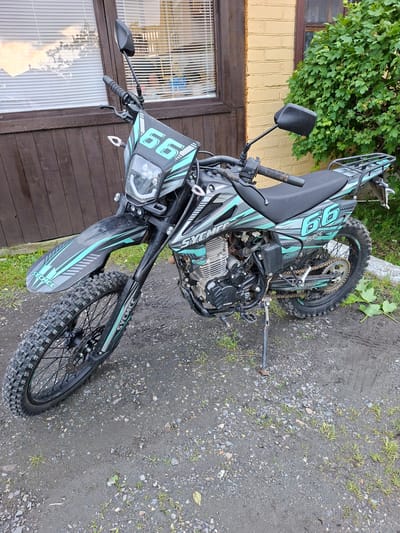 A black and teal dirt bike, identified by the "66" graphics and "SYMC" branding, is parked on a dirt and gravel surface. The motorcycle is positioned at an angle, with its front wheel turned slightly to the left. It is standing upright, supported by its kickstand. The bike features knobby tires suitable for off-road use and a rear rack.

In the background, a rustic wooden building with dark brown siding and a window covered by white blinds is visible. Adjacent to the building is a yellow brick structure. A dense green bush is growing to the right of the building. The overall environment appears to be an outdoor, possibly semi-rural or residential area. The ground is uneven, with patches of mud, small stones, and sparse blades of grass. There is no indication of human presence or activity in the image. The scene does not provide specific details that would allow for a precise location within the city of Pervouralsk, Russia.