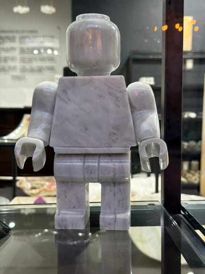 The image features a marble sculpture resembling a Lego figure, displayed inside a glass case. The sculpture is white with grey veining, characteristic of marble. The background is blurred, showing other objects within the case and the environment outside the display, suggesting a retail or museum setting. The location is Domingo Arenas, Mexico.