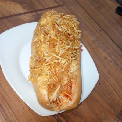 A single, elongated food item, resembling a loaded hot dog or a similar bread-based sandwich, is centrally positioned on a white, slightly rectangular plate. The bun is light golden-brown and appears soft, with a lengthwise cut revealing the interior. The filling consists of a creamy, pale yellow sauce, possibly a cheese or mayonnaise-based dressing, layered beneath a dense covering of golden-brown, thinly shredded, crispy fried potatoes (shoestring fries or *batata palha*). Near the bottom right end of the food item, a small amount of red sauce, consistent with ketchup, is visible within the filling. The white plate, which exhibits a subtle red decorative motif on its right edge, rests on a wooden surface. This surface is composed of multiple light to medium brown wooden planks, arranged diagonally, displaying distinct grain patterns. No individuals or specific identifying environmental markers related to the city of El Limón, Venezuela, are discernible within the image.