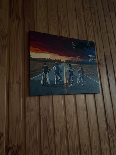The image displays a rectangular canvas art piece depicting four children on bicycles riding down a road. The artwork has a dark, stormy sky with hints of lightning and a dramatic sunset. A "Welcome to Hawkins" sign is visible on the right side of the road. The canvas is mounted on a wall covered in vertical wooden planks with a visible grain pattern. The scene is static and illustrative, with no discernible action or interaction occurring. The artwork's style is stylized and graphic. The location of this image is not determinable from the visual information provided.