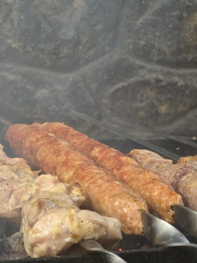 This media file shows a close-up, top-down view of meat grilling on skewers over a dark, textured surface, possibly a grill or fire pit. The main subjects are several pieces of meat, including two large sausages and pieces of what appear to be chicken. The sausages are elongated and have a reddish-brown, slightly wrinkled skin, indicating they are cooked or cooking. They are seasoned with black pepper. The chicken pieces are paler, with visible seasoning and some browning. Metal skewers hold the meat, and part of a metal tool, possibly tongs, is visible, interacting with the sausages. Smoke is present, partially obscuring the background and adding to the impression of a cooking process. The background is a dark, out-of-focus texture, which could be a grill grate or the surrounding environment. There is no visible text in the image. The scene suggests an outdoor cooking event, likely a barbecue or grilling session. The lighting is not indicative of a specific time of day, and there are no weather cues. Given the location context of Pidhorodne, Ukraine, this could be a common outdoor meal preparation. The overall impression is of food being cooked, with a focus on the meat and the grilling process.