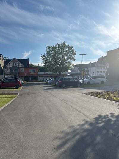 The image depicts a sunlit parking lot in the city of Waldbröl, Germany. In the foreground, a paved asphalt surface extends into the distance, with faint tire marks visible. To the left, a neatly mown lawn borders the parking area, separated by a concrete curb.
Several vehicles are parked in the lot. A red hatchback is parked on the left, facing away from the viewer. Behind it, a line of cars is parked, including a black SUV, several silver and gray sedans, and a white van.
In the background, a row of buildings lines the street. The building on the far left features a traditional half-timbered facade. Adjacent to it is a red building with a sign that reads "Strohkuhle" and another sign in German that likely indicates a business. Further down the street, other buildings are visible, including a multi-story apartment building. A prominent tree stands in front of these buildings, its branches casting shadows on the parking lot. The sky above is a clear blue, with wispy white clouds and bright sunlight suggesting a pleasant day. A lamppost is also visible in the scene.
