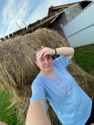 The image shows a young woman in a blue t-shirt, taking a selfie outdoors. She is standing in front of a large haystack, with a dilapidated building and a metal fence in the background. The sky above is partly cloudy. The woman has her hand on her forehead, possibly to shield her eyes from the sun. The setting appears to be a rural area, possibly on a farm. There is no identifiable information in the image to confirm the location as Балдейка, Russia.