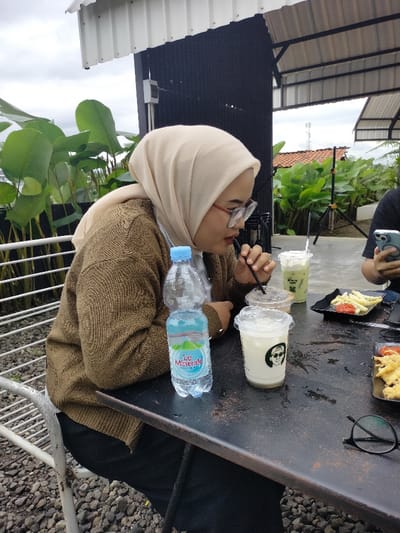 The image depicts an outdoor dining scene, likely at a cafe or restaurant in Dramaga, Indonesia, under an overcast sky. A woman, the primary subject, is seated at a dark, rustic table. She is wearing a light beige hijab, a brown knitted cardigan, and eyeglasses, and is actively drinking from a clear plastic cup with a black straw.
On the table, a clear plastic bottle of "Le Minerale" water is visible. Alongside it are several drinks in clear plastic cups: one with a white beverage featuring a stylized face logo, another with a brown liquid, and a taller cup containing a green beverage topped with whipped cream, possibly a matcha latte. Two black plates hold snacks, one with french fries and red sauce, and another with fried food. A pair of black-rimmed glasses also rests on the table. Part of another person is visible on the right, holding a smartphone.
The setting includes white metal chairs and a ground covered with small stones. The background features lush green tropical plants with large leaves. Behind these, modern-industrial structures with black corrugated walls and white metal roofs are visible, along with a distant red-tiled roof. The scene suggests a relaxed social gathering enjoying drinks and snacks.