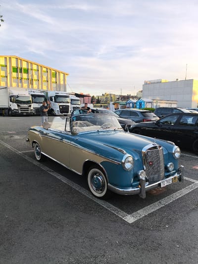 An outdoor, daytime, slightly overcast scene shows a parking lot near a building in Romanshorn, Switzerland. A vintage, two-tone blue and beige Mercedes-Benz convertible, license plate TG-7932, is parked in the foreground. The car has its top down and a fluffy object, possibly a blanket, is visible on the passenger seat.

Two men are standing near the car. One man, wearing a grey t-shirt and shorts, is standing to the left of the car and waving. The second man, wearing a dark t-shirt, is sitting in the driver's seat of a black car parked behind the convertible. Several white trucks are parked in the background, along with other cars and a light yellow building with green window shutters. The parking lot pavement is dark grey asphalt. The sky is a pale blue with wispy white clouds.