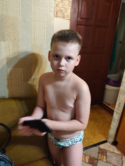 A young boy, appearing to be around five years old, stands shirtless and wearing patterned underwear. He has short, light brown hair and a serious expression on his face. The boy is holding a black object in his hands, which are blurred due to motion.
The boy is indoors, in what appears to be a domestic setting. The background includes a wall with textured wallpaper featuring a neutral, striped pattern. To the right of the boy, there is a dark wooden door. Further in the background, a white bucket with a purple lid and another white bucket are visible, suggesting a utility area or bathroom. The flooring is a mix of patterned tiles and what appears to be linoleum. A portion of a brown upholstered couch is visible on the left side of the image, with a black bag resting on it.
The lighting suggests it is daytime, possibly late afternoon or early evening, given the shadow cast on the wall behind the boy. The overall atmosphere is casual and candid, capturing a moment with the child. There is no visible text in the image. The location is identified as Bor, Russia.