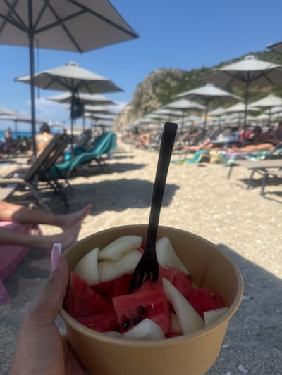 A person is holding a container of watermelon and melon pieces with a black plastic fork inserted. The background suggests a beach in the Municipal Unit of Lefkada, Greece, with sun umbrellas, beachgoers lounging on beach chairs, and a clear blue sky. The person's fingernails are painted with a French manicure. The scene is in daylight, likely during summer.