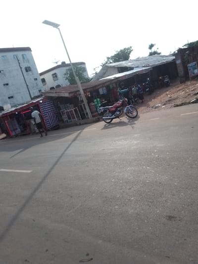 The image depicts a street scene in Orlu, Nigeria. A red motorcycle is parked on the asphalt road. Adjacent to the road are makeshift shops under corrugated metal roofs. Several people are present in the shops and along the roadside. A man in a white shirt is walking away from the shops. In the background, a taller, multi-story building stands beside a smaller building with a satellite dish on its roof. A streetlight pole is also visible.