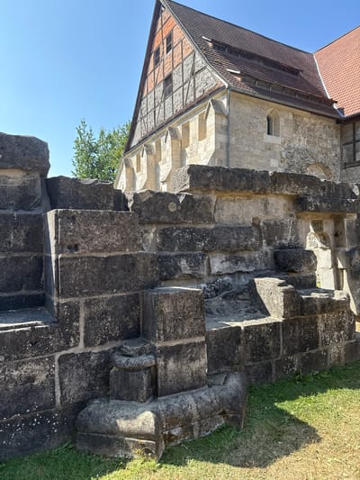 The image shows the exterior of a historic building, likely a church or monastery, in Walkenried, Germany. In the foreground, there is a weathered stone wall or ruin constructed from large, dark gray blocks. The blocks vary in size and are stacked unevenly, suggesting age and perhaps reconstruction. Some of the stones have visible lichen or moss growth. To the left of the stone structure, there is a patch of green grass.

Behind the stone structure, a portion of a building with a red-tiled roof is visible. The upper part of this building features a steeply pitched gable with exposed timber framing and brick infill, a style indicative of traditional German architecture. The lower part of the building is constructed from lighter colored stone blocks. The building has a large, prominent roof with dormer windows featuring louvers.

A clear blue sky dominates the background, with a few faint, dark specks that could be distant birds or dust motes. A green tree with lush foliage is partially visible behind the stone structure and to the left of the building. The sunlight suggests it is daytime, and the shadows indicate a sunny environment. There are no people visible in the image. The overall scene depicts a historical architectural site under a clear sky.