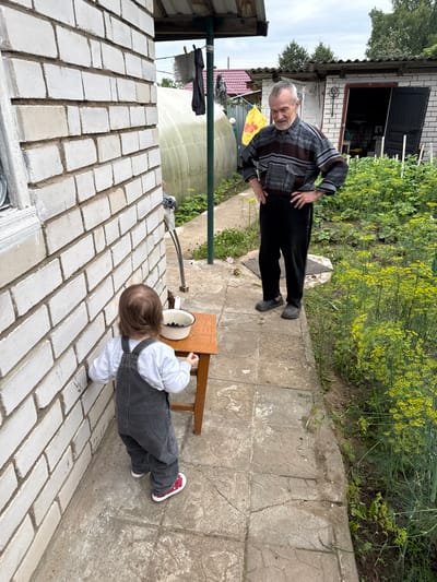 In the city of Vyshegor, Russia, an elderly man with grey hair and a patterned shirt stands with his hands on his hips, facing a young child. The child, wearing a grey jumpsuit and red-and-white polka dot shoes, stands with their back to the camera, looking at a white bowl filled with dark berries on a small wooden table. The scene takes place outdoors, next to a white brick building. A greenhouse is visible in the background, along with a small shed with an open door. Lush green plants, including dill, surround the area, suggesting a garden setting. The ground is paved with concrete slabs. The sky is overcast.