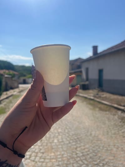A person's hand, with painted fingernails, holds a white paper cup against a bright blue sky. In the background, a cobblestone street leads through a village. The architecture appears to be traditional Bulgarian, with low-rise buildings, some with grey and white facades, and terracotta roof tiles visible. A prominent brick chimney stands against the sky. The overall scene suggests a sunny day in the village of Zelenikovo, Bulgaria, possibly during a tourist visit. The cup shows some text, but it is illegible in the image.