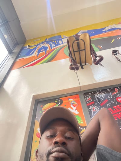 A man wearing a brown cap is taking a selfie. He is indoors, possibly in a restaurant or eatery, as there are fast food-themed decorations on the wall, including images of fries, burgers, and tacos. A child's horse saddle toy is also attached to the wall. The environment includes a window with daylight shining through. It is not possible to confirm the location of Rigachikun, Nigeria based on visual features present in this picture.