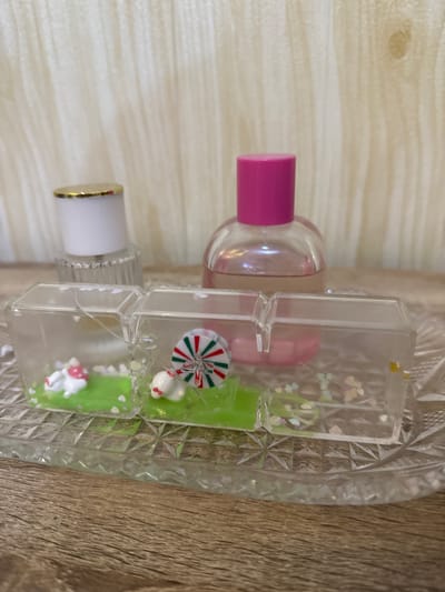 The image shows a close-up of a decorative arrangement on a wooden surface, set against a textured, light-colored background. The central focus is a clear, multi-compartment plastic display case containing miniature scenes. In the leftmost compartment, a small white figure with a red mushroom-shaped cap is positioned on a green base dotted with tiny white hearts. The middle compartment features a similar white figure, this time with a red ribbon around its neck, sitting on a green base adorned with a pinwheel-like decoration of red, green, and white stripes. The rightmost compartment contains only small, colorful heart-shaped confetti scattered on a clear base.
Behind the plastic case, two bottles are visible. The one on the left has a white cap with a gold rim and a clear, ridged glass body. The bottle on the right is clear with a bright pink cap and contains a pinkish liquid. The entire arrangement rests on a clear, ornate glass tray with a starburst pattern.
This image was taken in the city of Pematang Siantar, Indonesia.