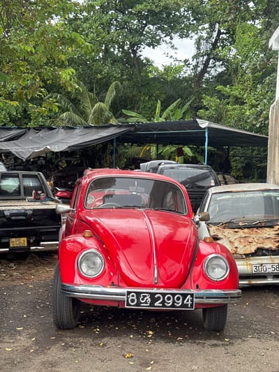 The image shows a red Volkswagen Beetle parked on a road in Sri Jayewardenepura Kotte, Sri Lanka. The car is facing the viewer, and its license plate is visible. Other vehicles are parked behind the Beetle. The scene is partially shaded by a canopy structure, with trees and foliage visible in the background.