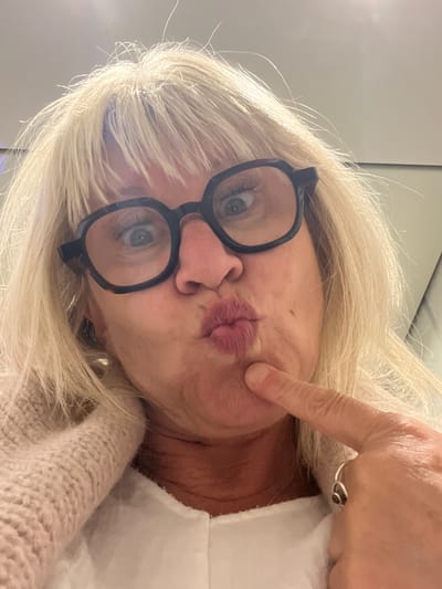 The image is a close-up, angled selfie of a person, likely a woman, taken in Palavas-les-Flots, France. The subject has long, blonde hair that frames her face and flows downwards towards the bottom of the frame. She is wearing black-rimmed glasses, through which her blue-green eyes are visible, looking directly at the camera. Her lips are pursed in a "duck face" pose with pink lipstick, and her right index finger is playfully pressing into her left cheek, causing a slight indentation. A simple silver ring with a dark stone is visible on her ring finger. The person is wearing a light-colored, possibly knitted, garment, part of which is visible on the right side of the frame. The background appears to be a plain, light-colored wall or ceiling, suggesting an indoor setting. The overall expression and pose convey a playful and humorous mood. No text is visible in the image.