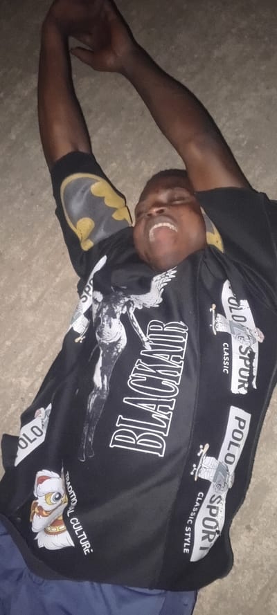A person lies on a grey surface, face upwards with their arms raised. The person is wearing a black t-shirt with various graphic designs, including a "BLACK AIR" print, "POLO" text, a Batman logo on the sleeve, and a lion figure. The person's mouth is open, revealing their teeth. The overall lighting is dim.
