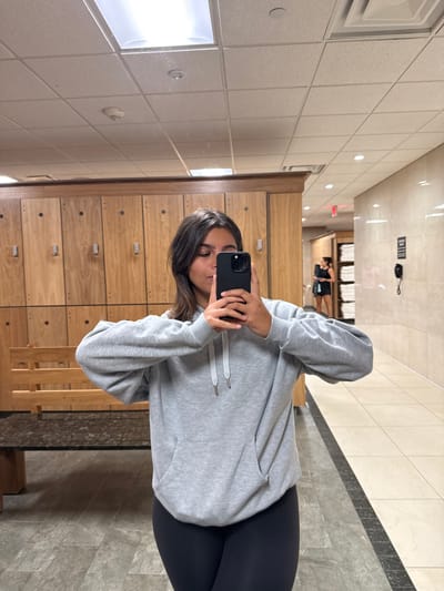 The image displays a full-body mirror selfie taken by a female subject in a locker room setting within a fitness or wellness facility, located in Plainsboro Township, United States. The subject, centered in the frame, has medium-length dark hair and is wearing a light grey hooded sweatshirt with white drawstrings and dark leggings. She holds a black smartphone horizontally with both hands, partially obscuring the lower half of her face as she photographs her reflection.

The environment behind her left shoulder features a row of light-toned wooden lockers with silver handles, beneath which is a wooden bench topped with a dark, possibly granite, surface. The floor consists of dark grey square tiles. The ceiling is a white suspended grid system, containing rectangular fluorescent light fixtures, smaller recessed lights, a circular smoke detector, and an air vent.

In the reflection to the right and in the background, a tiled hallway or open area is visible. The walls are covered in large, light beige tiles. A second female subject, dressed in dark clothing and carrying a dark bag, is observed walking away in the mid-ground of the reflection. Further back in the reflection, a black wall-mounted landline phone, a black sign with indistinct white text, and a red exit sign are visible on the tiled wall. Stacks of white folded towels are also discernible on shelving in the background reflection, consistent with a gym or spa amenity.