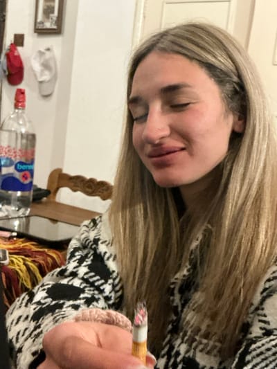 In El Rodeo, Argentina, a young person with long, blonde hair is shown indoors, close-up. The person is wearing a black and white patterned jacket and has their eyes closed, with a slight smile on their face. They are holding a lit cigarette in their right hand, which is in the foreground of the image. To the left, a bottle of water with a red and blue label is visible on a reflective surface, possibly a table. In the background, a wooden chair with a carved design can be seen, and on the wall, there are framed pictures and two hats hanging. The lighting suggests an indoor setting, likely a home environment.