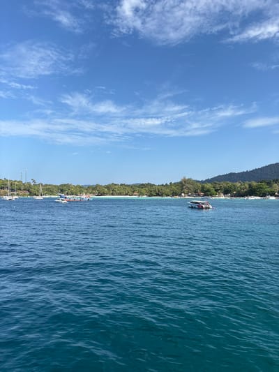 The scene depicts a clear-day panoramic view of the coastline of Ban Ko Li Pe, Thailand. The upper portion of the image shows a bright blue sky with scattered wispy white clouds. The lower two-thirds of the frame consists of calm, deep blue-green ocean water exhibiting minor surface ripples. The background features a white sandy beach fringed by dense tropical vegetation, including numerous palm trees and other foliage, which extends to a forested hillside on the right.
In the water, several vessels are visible:
* On the far left, multiple white-masted sailboats are anchored or moored near the shoreline.
* Closer to the center-left, a light-colored long-tail boat and another smaller vessel are present in the water near the beach.
* In the mid-right, a white and red trimaran-style speedboat, equipped with at least three outboard engines, is stationary on the water.
On the land, a tall antenna tower is discernible amongst the trees on the far left of the coastline. Several structures with reddish-brown roofs are partially visible through the vegetation along the beachfront. No individual persons or specific interactions are clearly identifiable within the scene.