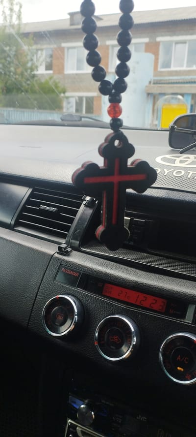 The image shows the interior of a car, focusing on the dashboard and a hanging car accessory. A black beaded string with a red and black cross pendant is prominently displayed, hanging from the rearview mirror area. The dashboard features various controls, including climate control knobs and a digital display showing "27C" for the outside temperature. A "PASSENGER" indicator light is also visible. In the background, through the car's windshield, a brick building with white-framed windows and some trees can be seen, suggesting an outdoor setting in a populated area. The overall scene is a snapshot from within a vehicle, likely parked or in slow-moving traffic.