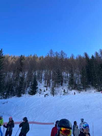 This media file captures a sunny winter day on a ski slope in Courmayeur, Italy. The image, rotated counter-clockwise, shows a clear, bright blue sky on the left side, indicating excellent weather. A dense line of trees, including both bare deciduous trees and dark green coniferous evergreens, runs horizontally across the middle of the frame, forming a natural border between the sky and the ski area.

On the right side of the image, a snow-covered ski slope is visible, featuring visible ski tracks on the white surface. Several skiers are present on the slope, dressed in full ski gear, including helmets, goggles, and winter jackets. In the foreground, a skier wears a black helmet with a striking yellow/orange mirrored visor and a red jacket; a "Dynastar" logo is faintly visible on their helmet. Directly behind them, another skier wears a white "POC" helmet with blue mirrored goggles and has a GoPro camera mounted on top. Further down the slope, additional skiers can be seen, dressed in various colored jackets (dark green, black/yellow, light green), holding ski poles. They appear to be either queuing or gathered at a starting point on the slope. A red safety net or rope runs along the far right edge of the ski slope. The bright sunlight suggests it is likely midday or early afternoon.