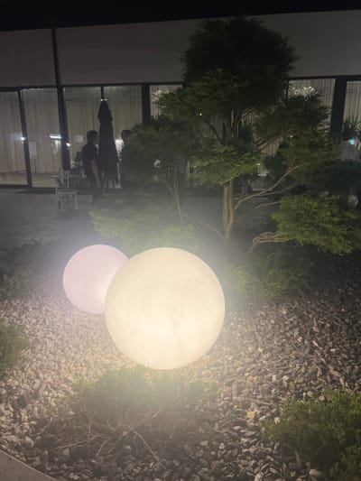 In Achadara, Abkhazia, at night, two luminous spherical objects, likely outdoor lamps, are prominently displayed on a ground covered with gravel and low-lying green vegetation. The larger sphere, on the right, emits a bright, warm white light, while the smaller sphere to its left emits a softer, pinkish-white glow. Behind these spheres, a dark, well-maintained tree with intricate branching stands. In the background, through a large glass facade, several individuals are visible. Two men, one in a black shirt and the other with his back to the camera, appear to be standing near tables and chairs, suggesting an outdoor seating area of a restaurant or establishment. The overall scene suggests an outdoor evening setting, illuminated by decorative lighting.