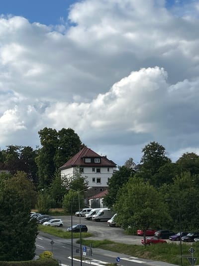 In Waldbröl, Germany, a residential building with a red tiled roof is visible amidst a dense green canopy of trees. In front of the building, a parking lot contains several vehicles, including white vans, a white hatchback, and a dark sedan. Adjacent to the parking lot is a road with white lane markings, where a dark sedan is parked. The sky above is filled with dramatic, billowing clouds against a blue backdrop.