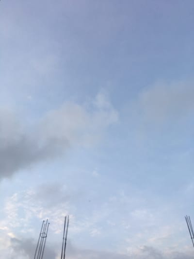 The scene, located in Agunmo, Nigeria, features a partially cloudy sky. The upper and central areas display a light blue sky interspersed with diffuse white clouds. Towards the lower left, denser grey cloud formations are present. The extreme bottom edge of the image reveals the upper portions of three separate vertical rebar bundles, consistent with structural elements of a building under construction. No people, actions, or interactions are discernible.