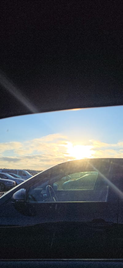 The image is a photograph taken from inside a vehicle looking out at a parking lot with a bright sun setting or rising in the sky. The foreground shows the interior of a car, with the dashboard and a portion of the driver's side window visible. Outside the window, several cars are parked in rows under a sky filled with clouds. The sun is a bright, intense light source, casting rays across the scene. The overall lighting suggests it is either early morning or late evening. The location in Neustadt-Glewe, Germany, is not visually discernible from the image itself.