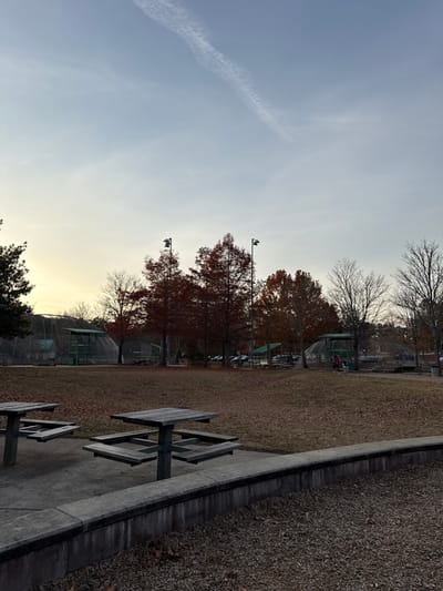 A landscape shows a playground and picnic area under a sky with a contrail. Tennis courts with nets are visible in the background, adjacent to autumn trees with changing leaves and light posts. Picnic tables stand on a concrete slab bordered by a bed of mulch. The trees have mostly bare branches at the top, suggesting a transitional season. The light indicates it's either early morning or late afternoon.