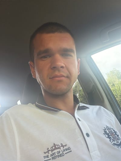 A male individual, estimated to be between 20 and 30 years old, with a short buzz cut and light facial stubble, is photographed in a selfie inside a vehicle. He is wearing a white pique polo shirt with a dark blue collar accented by thin red and white stripes. The shirt features two distinct embroidered emblems: a smaller dark design with a crown and the text "THE ART OF LIVING REFINED" on the left chest, and a larger, elaborate dark blue and red crest on the right chest. The car's interior, including a light grey headliner and a dark seat headrest, is visible behind him. Intense sunlight illuminates the scene from the right, creating highlights on the subject's face and the car's interior. Through the car's right-side window, bright green foliage and a clear sky are discernible. The subject maintains a neutral facial expression, and a small reddish mark is present on his forehead. The image captures a moment within a vehicle in Giurgiu, Romania.