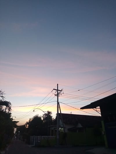 The image shows a street scene in Bungur, Indonesia at dusk. The sky transitions from blue at the top to pink and orange near the horizon. A utility pole stands in the foreground with several electrical wires extending across the frame. Houses and trees line the street, silhouetted against the colorful sky. One house has a visible tile roof. A street lamp is visible in the distance along the road. The street itself is dark, suggesting it is unlit. No people are immediately visible, though there may be a distant figure walking in the street.
