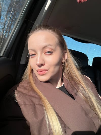 This image depicts a single adult female seated within the passenger compartment of a vehicle, presumed to be a car, in Spas-Zaulok, Russia. The woman, with a fair complexion, long blonde hair, and light-colored eyes, faces forward with a slight smile and direct gaze. She is wearing a dark brown coat over a dark undergarment and a single gold-colored, elongated link earring is visible on her right ear. Strong natural sunlight enters the vehicle from the left, illuminating her and casting shadows across her face and jacket. Through the left side window, bare trees with thin trunks are visible against a bright blue sky. The vehicle's dark-colored interior elements, including a seat, a window frame, and a portion of the headliner, are discernible. A blurry, dark red decorative item is partially visible hanging from the interior roofline towards the rear. The rear window shows an unobstructed bright blue sky.